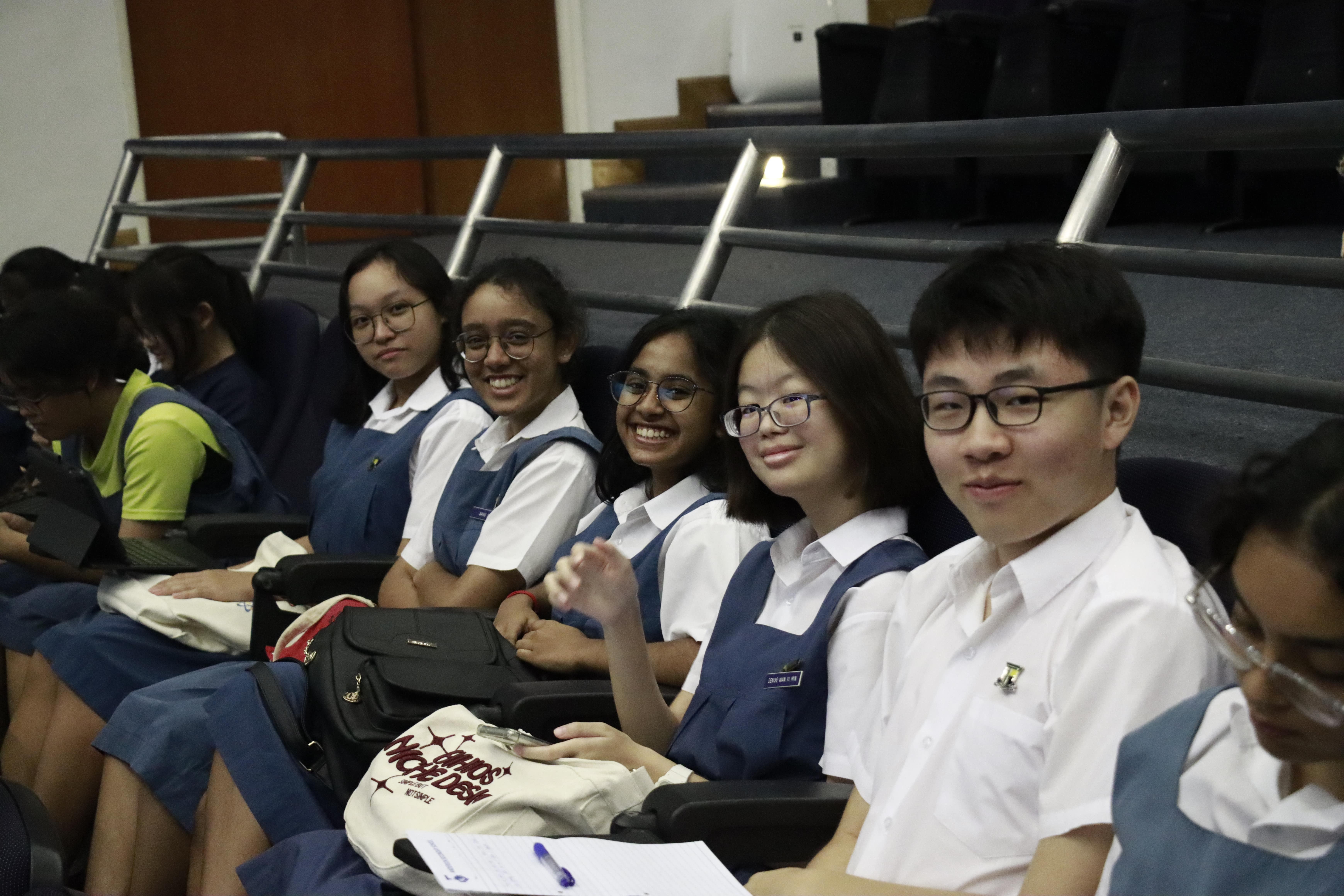 Several school students in blue and white uniforms sit in tiered auditorium seats, smiling toward the camera.